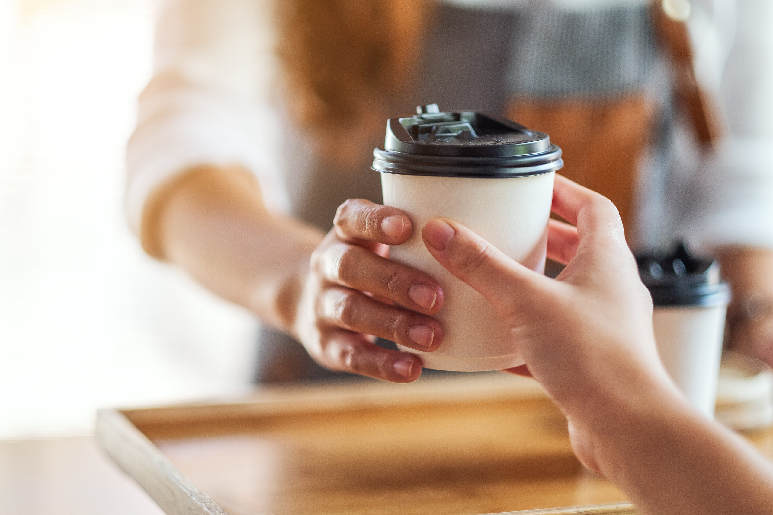 Barista überreicht einer Kundin bzw. einem Kunden einen weißen Coffee-to-go-Becher mit schwarzem Deckel.
