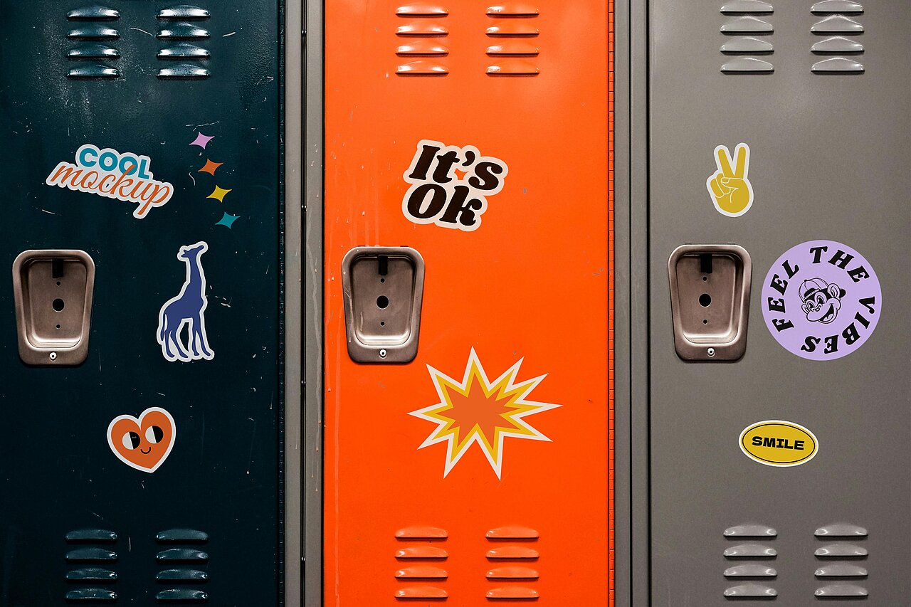 Mehrere farbige Sticker auf bunten Metallspinden, darunter Schriftzüge, Symbole und Illustrationen wie eine Giraffe, ein Peace-Zeichen und ein Smiley.