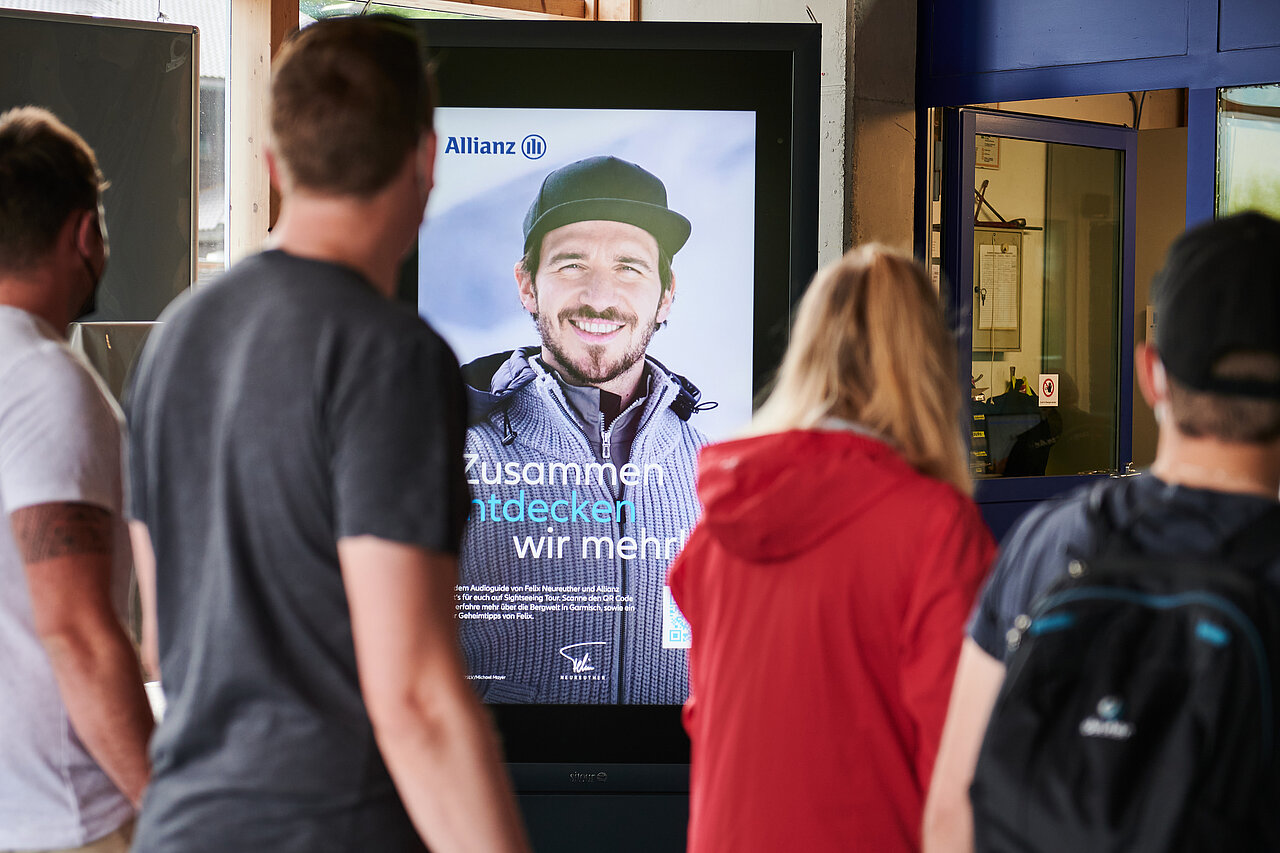 Digitale Werbetafel an einer Bergbahnstation mit Allianz-Werbung, die einen Outdoor-Sportler zeigt. Die Anzeige ist in einem modernen, hochauflösenden Display platziert.