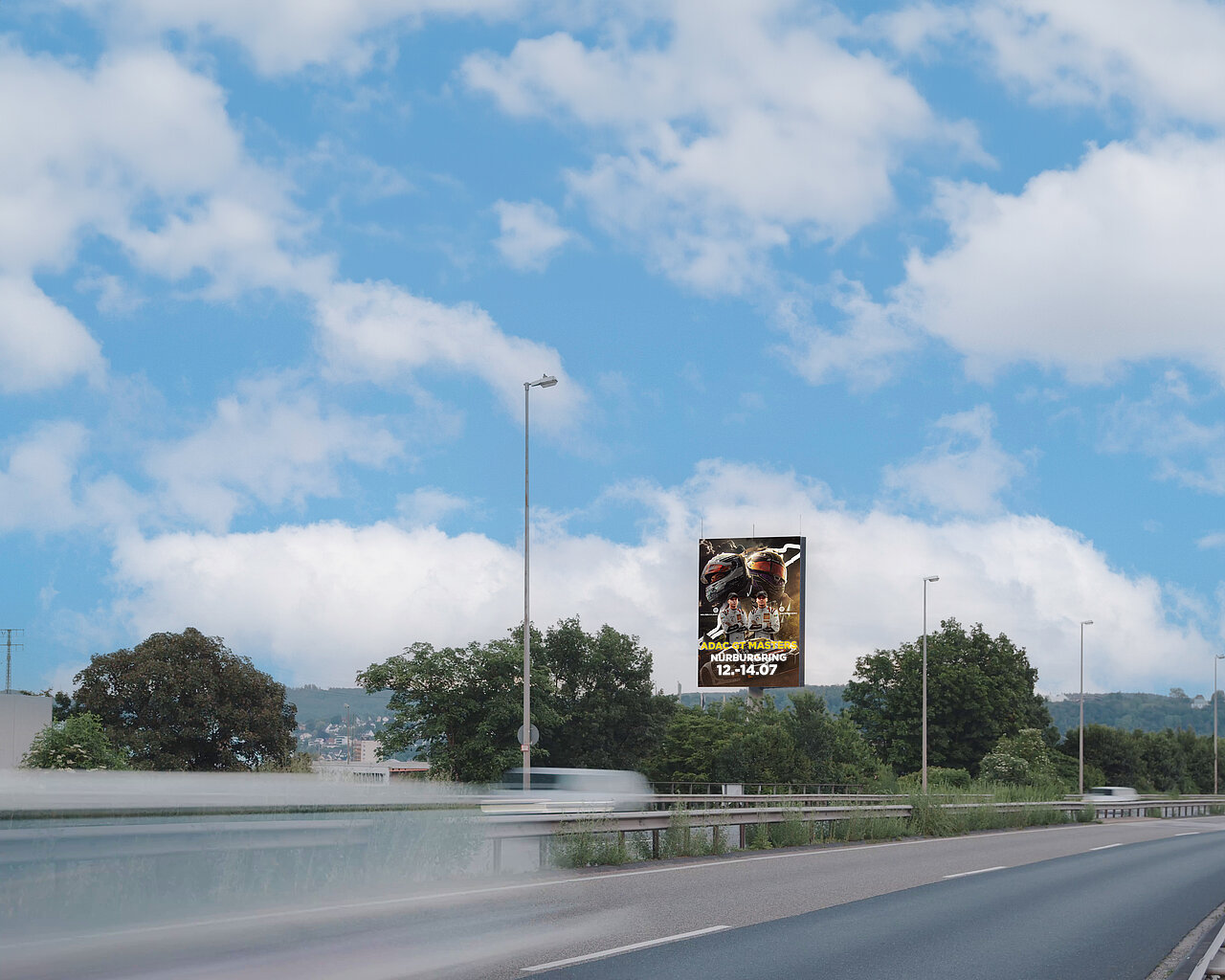 Digitaler Werbe-Tower an einer Straße mit Eventplakat für Nürburgring, vorbeifahrenden Autos und blauem Himmel mit Wolken