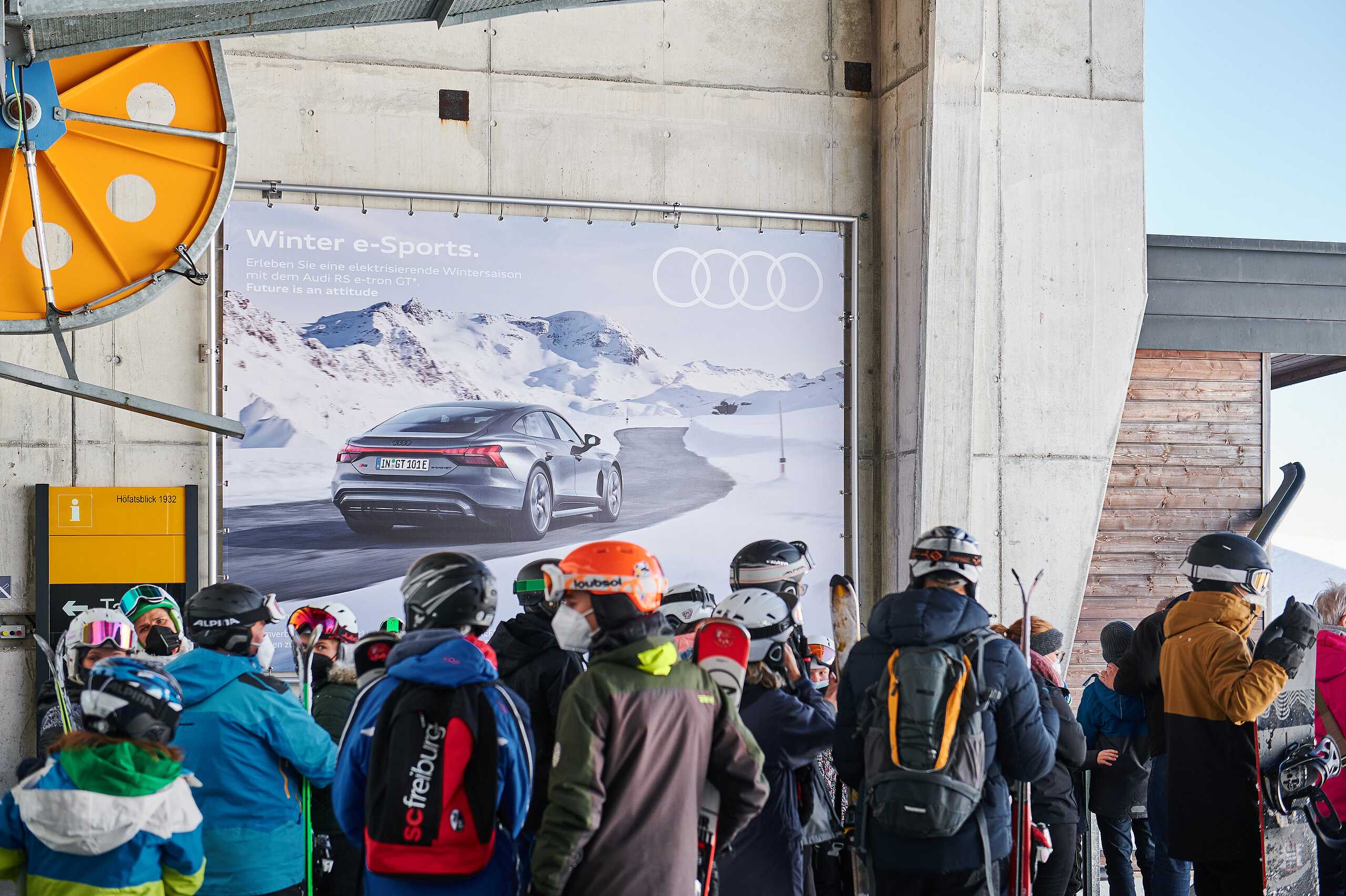 Megaposter von Audi mit Wintermotiv an einer Bergbahnstation, gesehen von wartenden Skifahrern in Skihelmen und Winterkleidung.