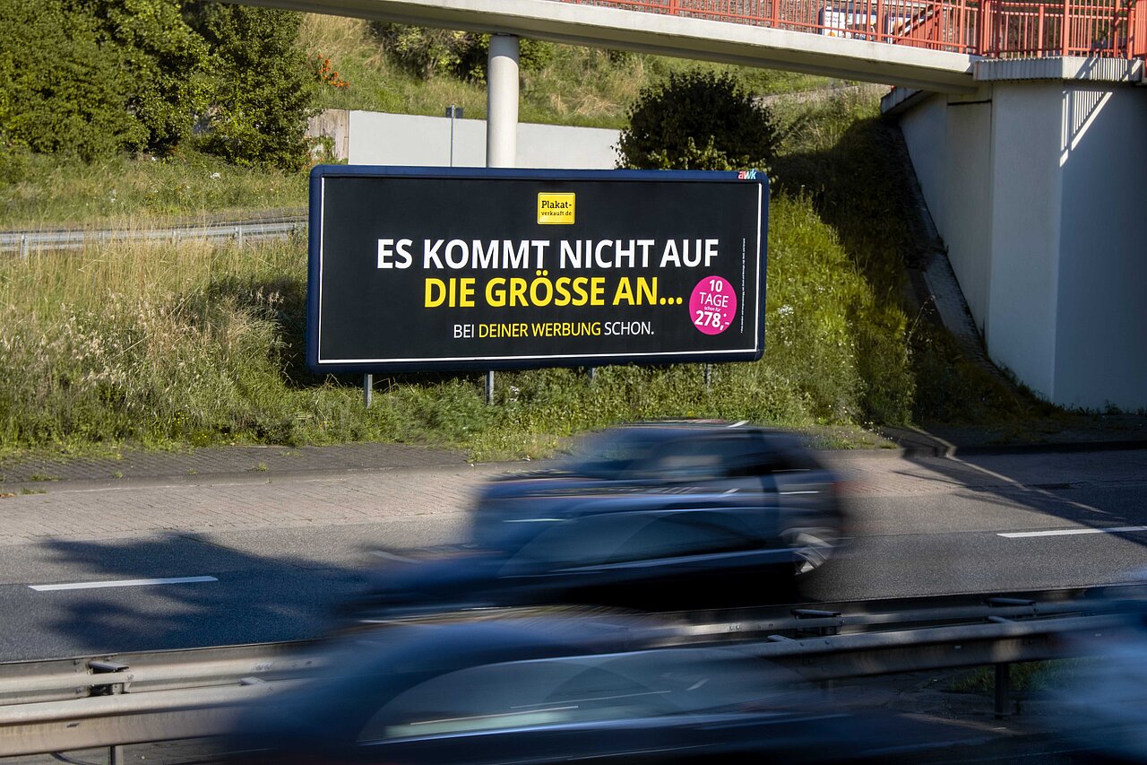 Megastar Plakatwand an einer stark frequentierten Straße mit dem Slogan 'ES KOMMT NICHT AUF DIE GRÖSSE AN' auf einem auffälligen Plakat. Optimale Platzierung für Werbewirkung im Verkehr.