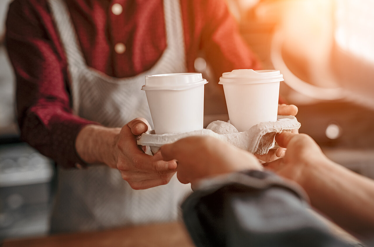 Barista überreicht zwei weiße Coffee-to-go-Becher in einem Getränkehalter an eine Kundin bzw. einen Kunden.
