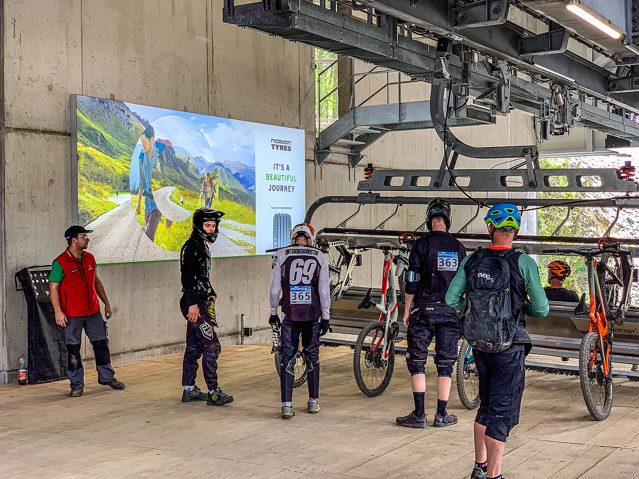 Digitale Werbetafel an einer Bergbahnstation, mit Werbung für Nokian Tyres, während Mountainbiker in Richtung Liftstation gehen.