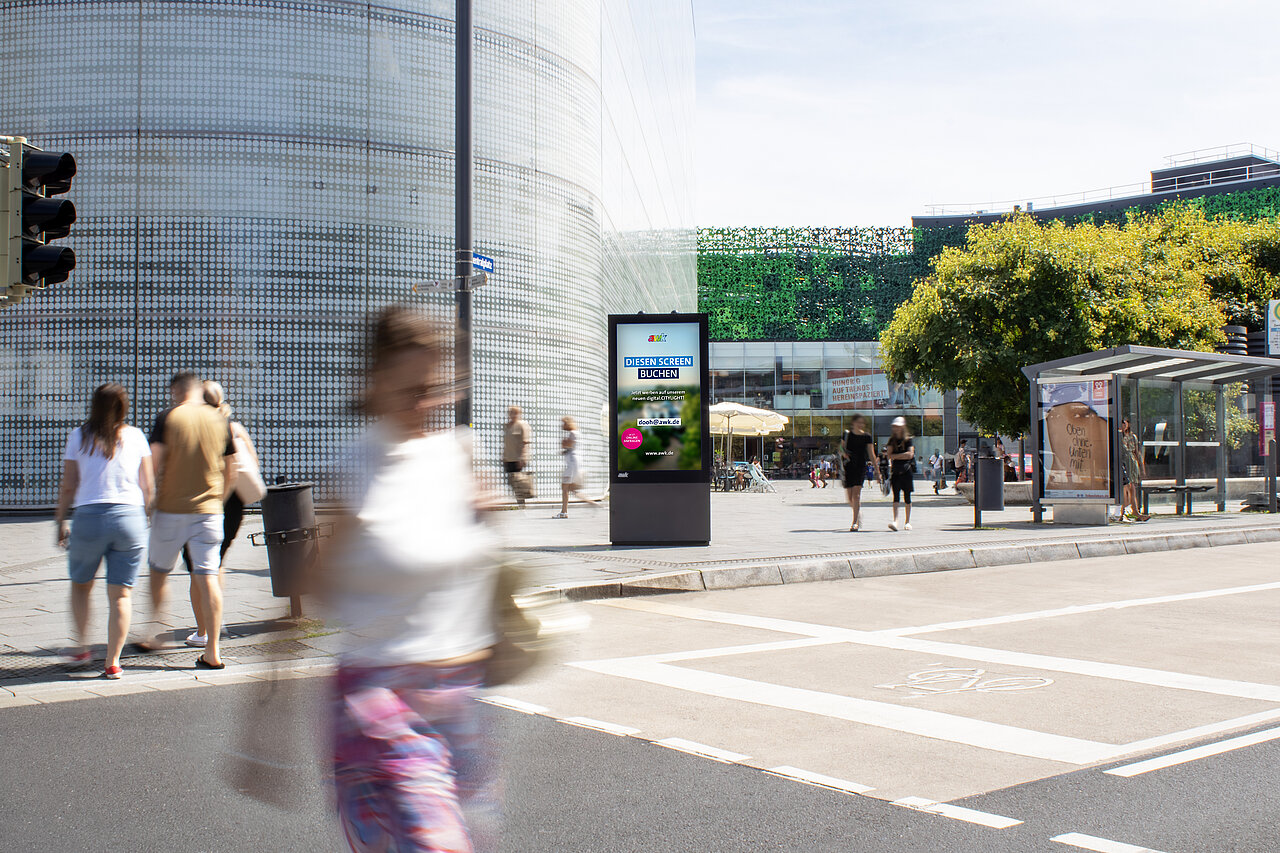 Digitales Citylight-Poster auf einem stark frequentierten Platz in der Innenstadt, umgeben von Fußgängern, moderner Architektur und einer nahen Bushaltestelle