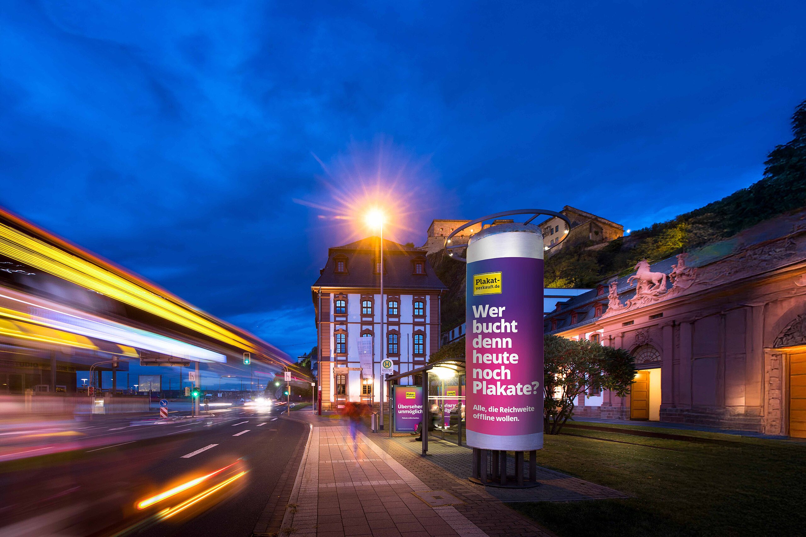 Plakatwerbung auf einer Ganzsäule von Plakat-verkauft.de bei Nacht, beleuchtet mit Verkehr auf der Straße.