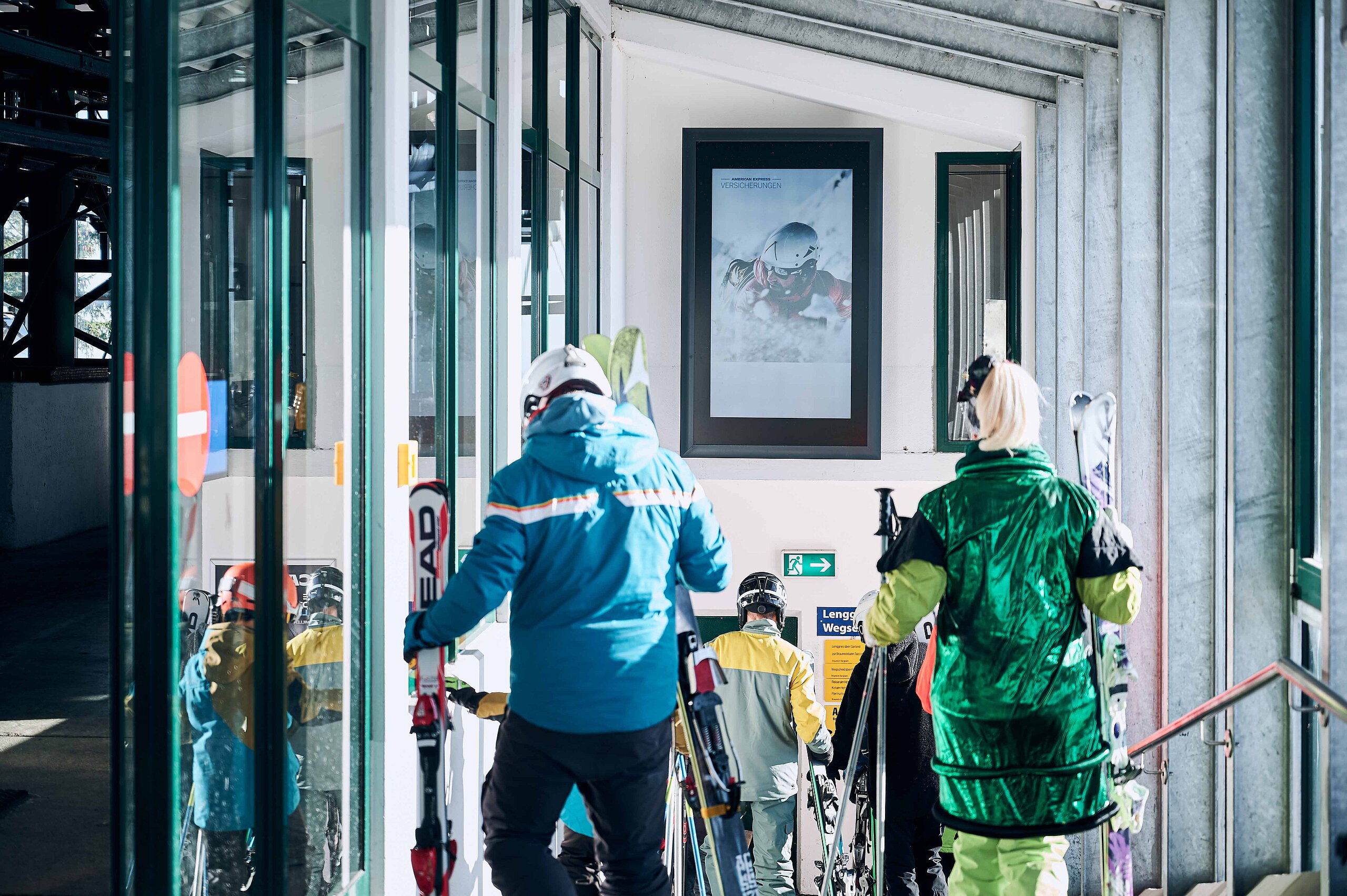 Digitale Werbetafel an einer Bergbahnstation, vor der Wintersportler in Skikleidung und mit Skiern vorbeigehen.