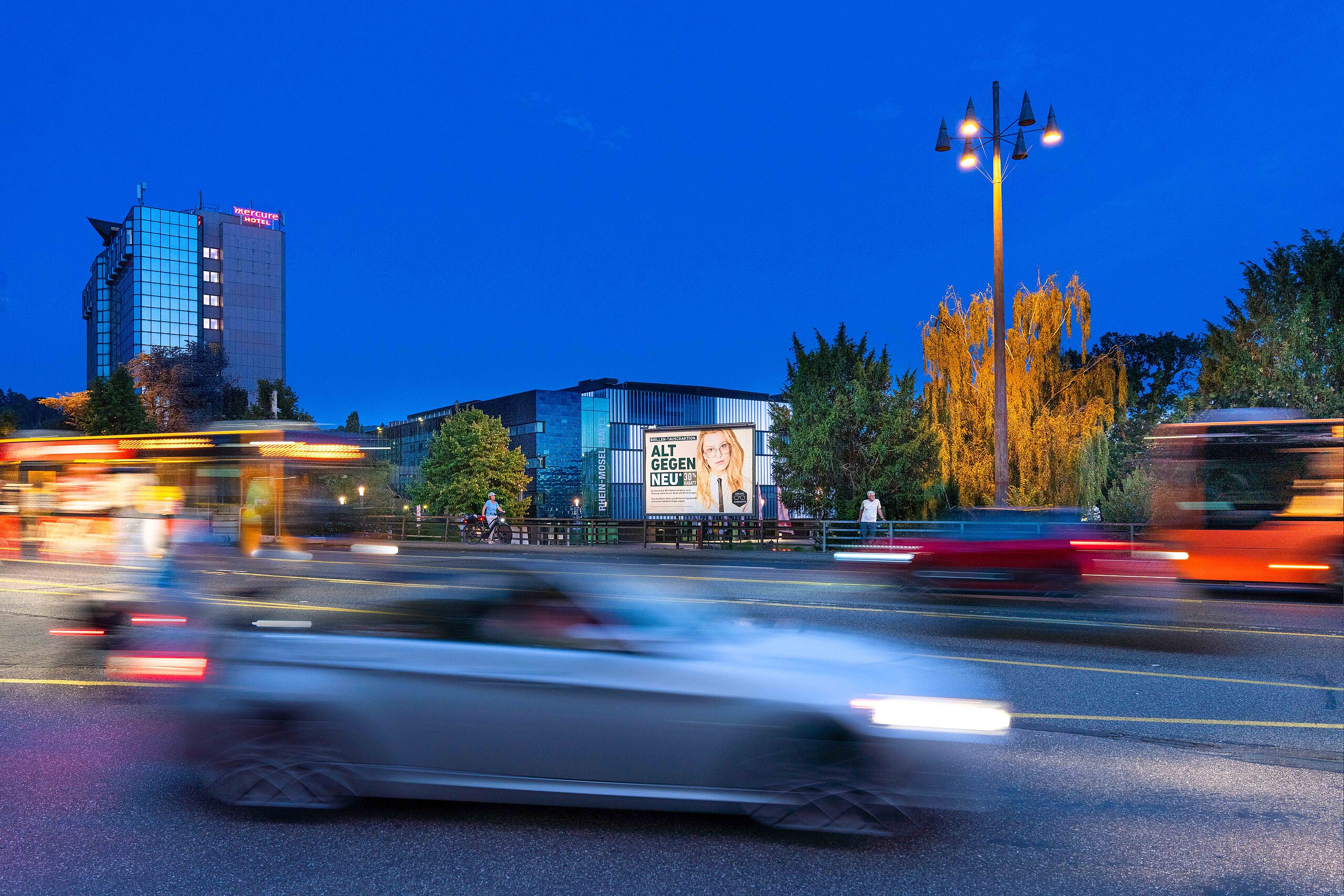 Beleuchtetes Großplakat mit Werbemotiv an einer stark befahrenen Straße bei Nacht, vorbeiziehende Autos und Busse – buchbar über Plakat-verkauft.de.