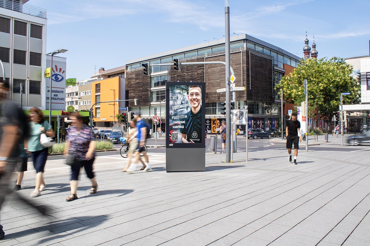 Digitale Vitrine am Zentralplatz zeigt ein Recruiting-Motiv mit einer lächelnden Person auf dem Screen, umgeben von vielen Passanten im Stadtzentrum.