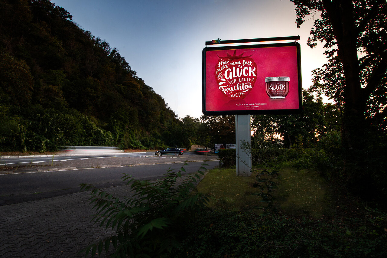 Werbeplakat mit dem Slogan 'Hier sieht man fast das Glück vor lauter Früchten nicht' und einem Glas Glück Marmelade. Das Plakat ist an einer gut sichtbaren Stelle entlang einer stark befahrenen Straße platziert.