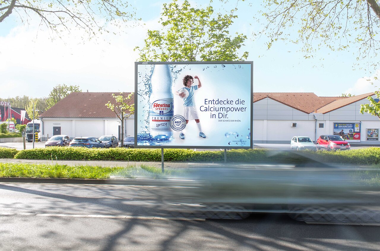 Werbeplakat für Fürstina Sprudel mit dem Slogan 'Entdecke die Calciumpower in Dir' und einem Kind, das eine Flasche Sprudelwasser hält. Das Plakat ist an einer gut sichtbaren Straßenecke platziert.