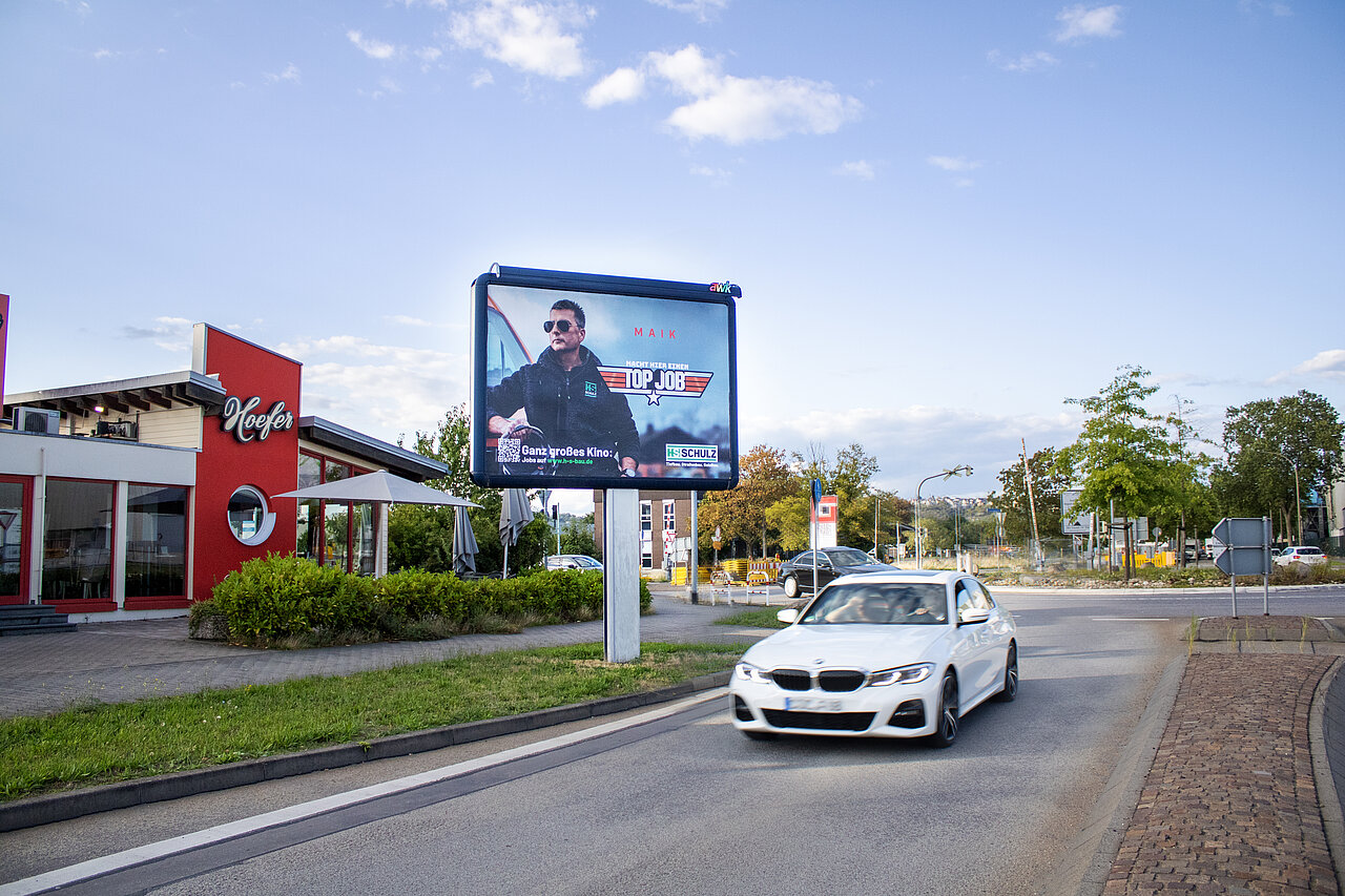 Großplakat mit Recruiting-Motiv an einer befahrenen Straße neben einem Restaurant, vorbeifahrende Autos – buchbar über Plakat-verkauft.de.