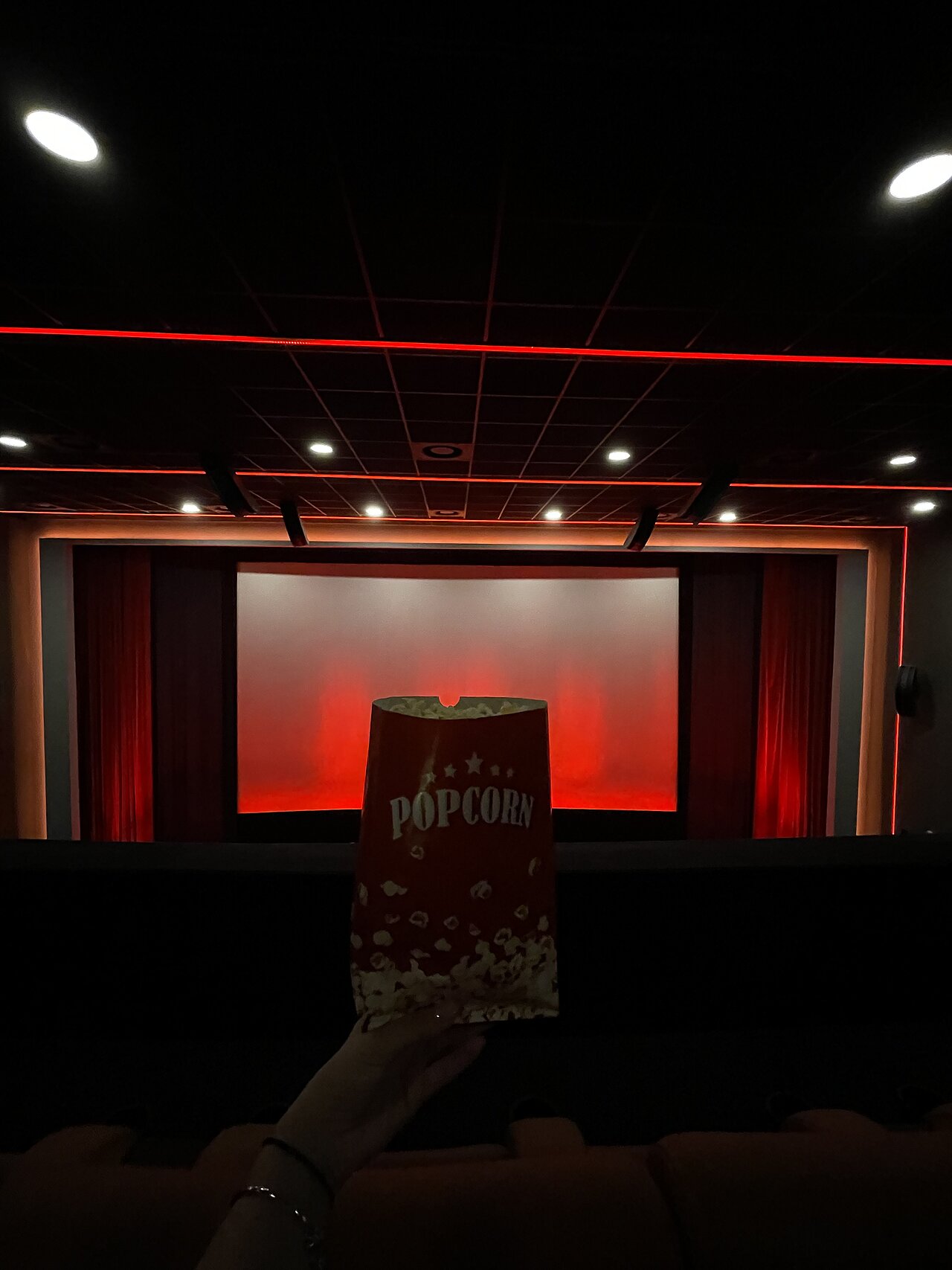 Kinosaal mit einem leeren Bildschirm, bereit für den Kinospot. Ein Popcorntütchen wird gehalten, während der Saal mit rotem Licht beleuchtet ist.