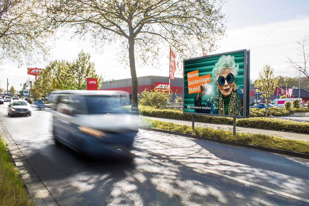 Werbeplakat mit dem Slogan 'Meine Geldanlage: Stabil!' und einer stilvollen Dame mit Sonnenbrille. Das Plakat ist an einer belebten Straßenecke platziert und sorgt für hohe Aufmerksamkeit im Straßenverkehr.