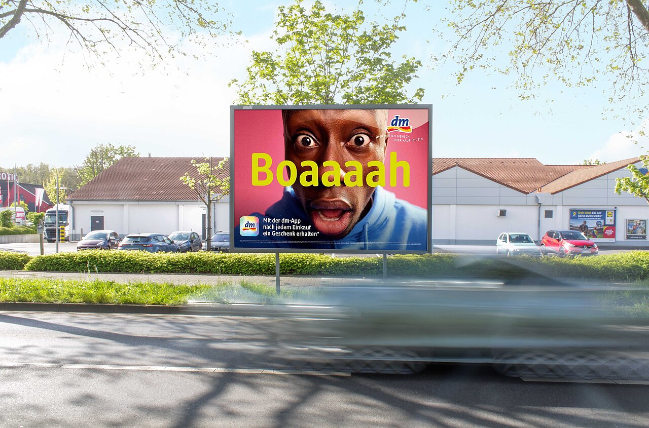 Großplakat an einer mehrspurigen Straße mit auffälligem „Boaaaah“-Motiv und vorbeifahrenden Autos vor einem Gebäudekomplex – buchbar über Plakat-verkauft.de.