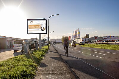 Plakat mit Aufschrift „Ihr Partner für Immobilien in Augsburg und Umgebung“ an Hauptstraße bei Sonnenuntergang