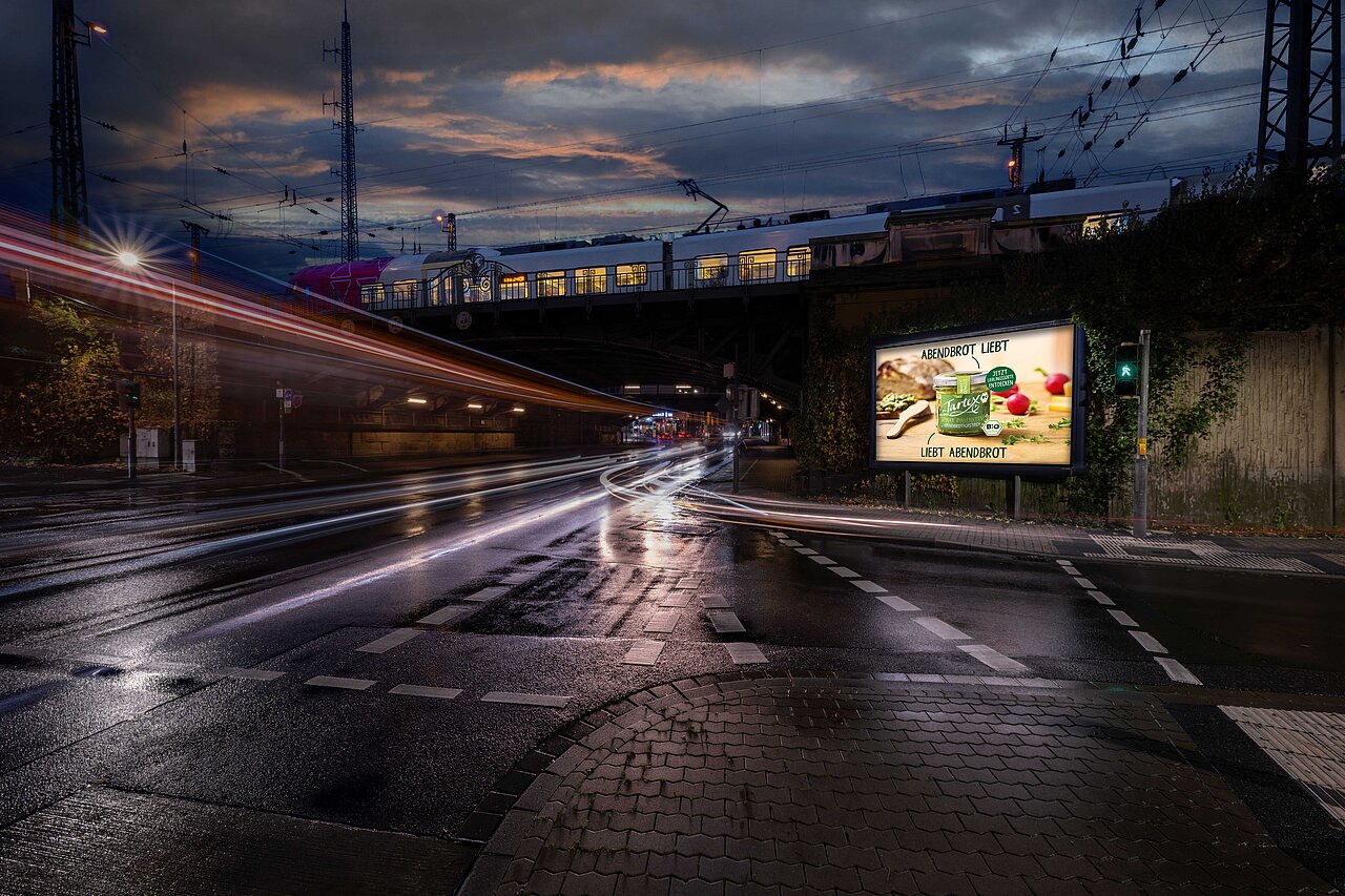 9-qm Werbeplakat an belebten Straßenkreuzung bei Dämmerung, mit einem vorbeifahrenden Zug und Straßenbeleuchtung im Hintergrund. Jetzt buchbar bei Plakat-verkauft.de.