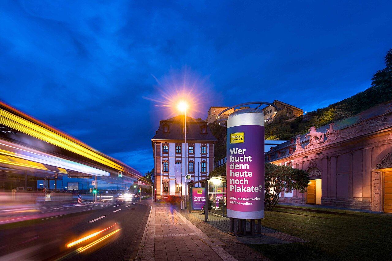 Ganzsäule mit Werbebotschaft von Plakat-verkauft.de bei Nacht, beleuchtet mit Verkehr auf der Straße und historischem Gebäude im Hintergrund.