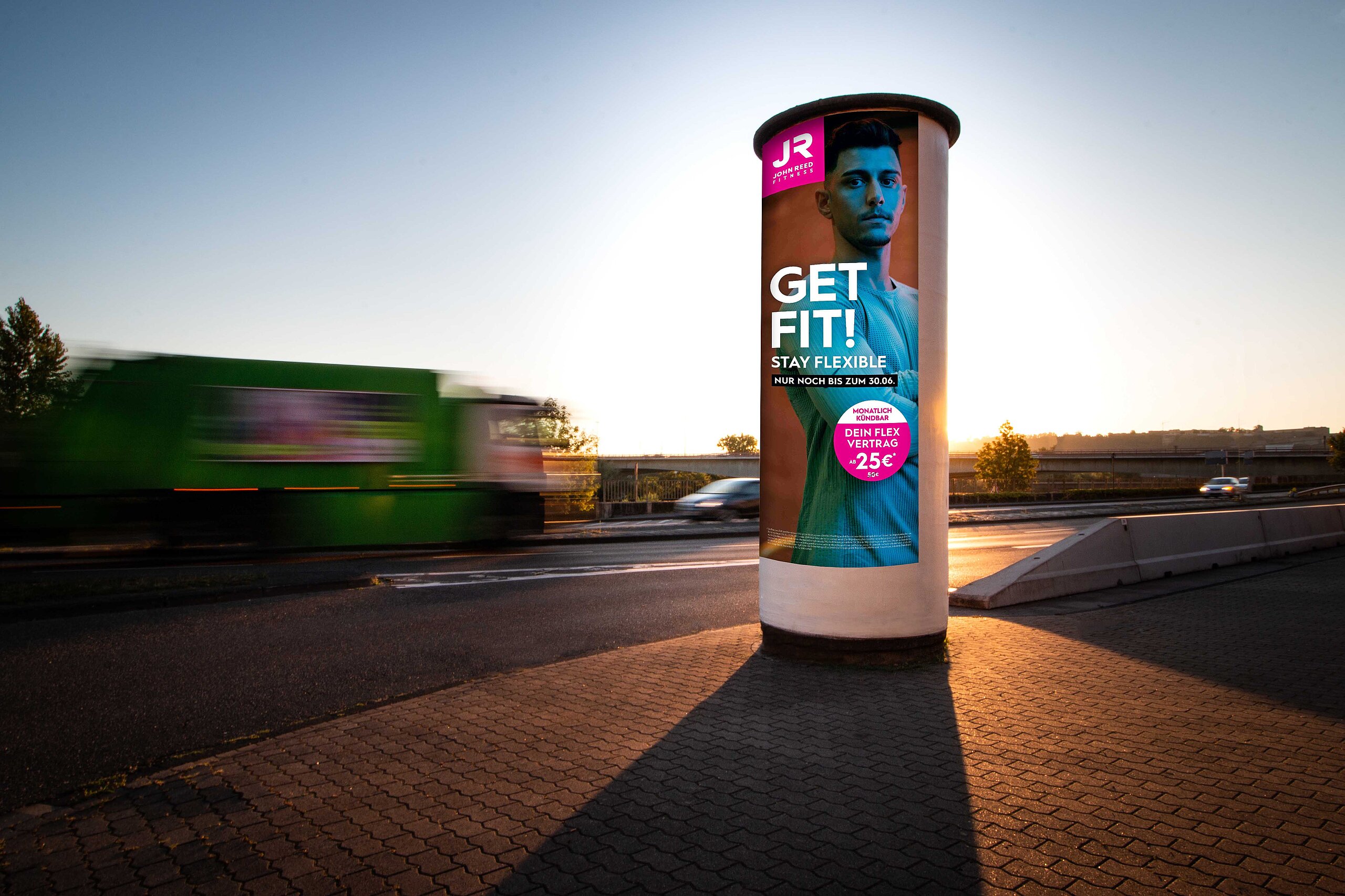 Ganzsäule mit Werbeanzeige für JR Fitness in der Abenddämmerung, umgeben von Fahrzeugen auf der Straße. Maximale Sichtbarkeit auf 360°-Fläche.