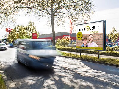 Plakat mit Aufschrift „Mein Optiker in Dieblich“ an befahrener Straße vor rotem Gebäude