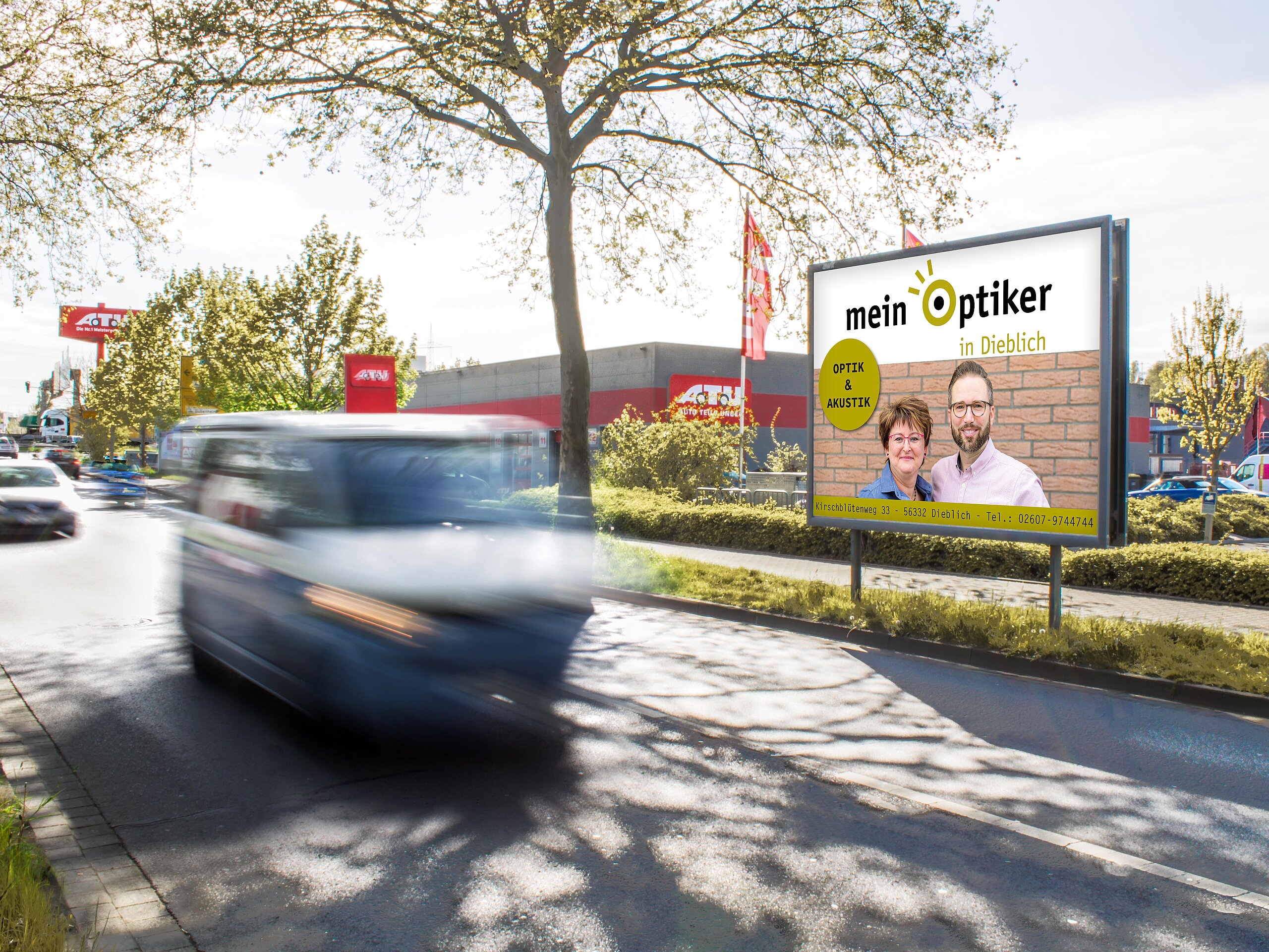 Plakat mit Aufschrift „Mein Optiker in Dieblich“ an befahrener Straße vor rotem Gebäude