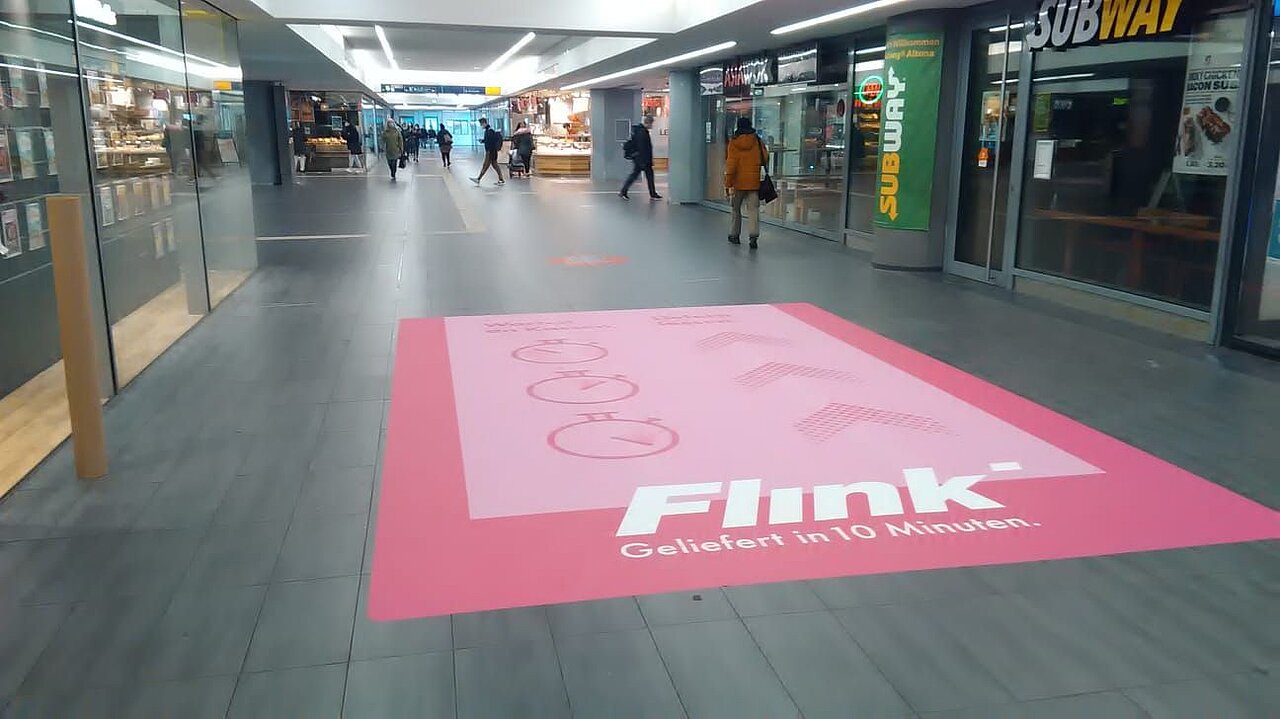 Pinkfarbene Flink-Floorgraphic im Einkaufszentrum, platziert im Laufbereich vor Geschäften und Passanten