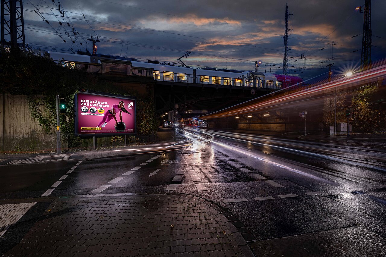 Plakat mit der Aufschrift 'BLEIB LÄNGER, ZAHL WENIGER!' zeigt eine sportliche Frau beim Training. Die Werbung ist an einer gut frequentierten Straßenkreuzung platziert und zieht die Aufmerksamkeit der Passanten und Autofahrer im Stadtverkehr an.