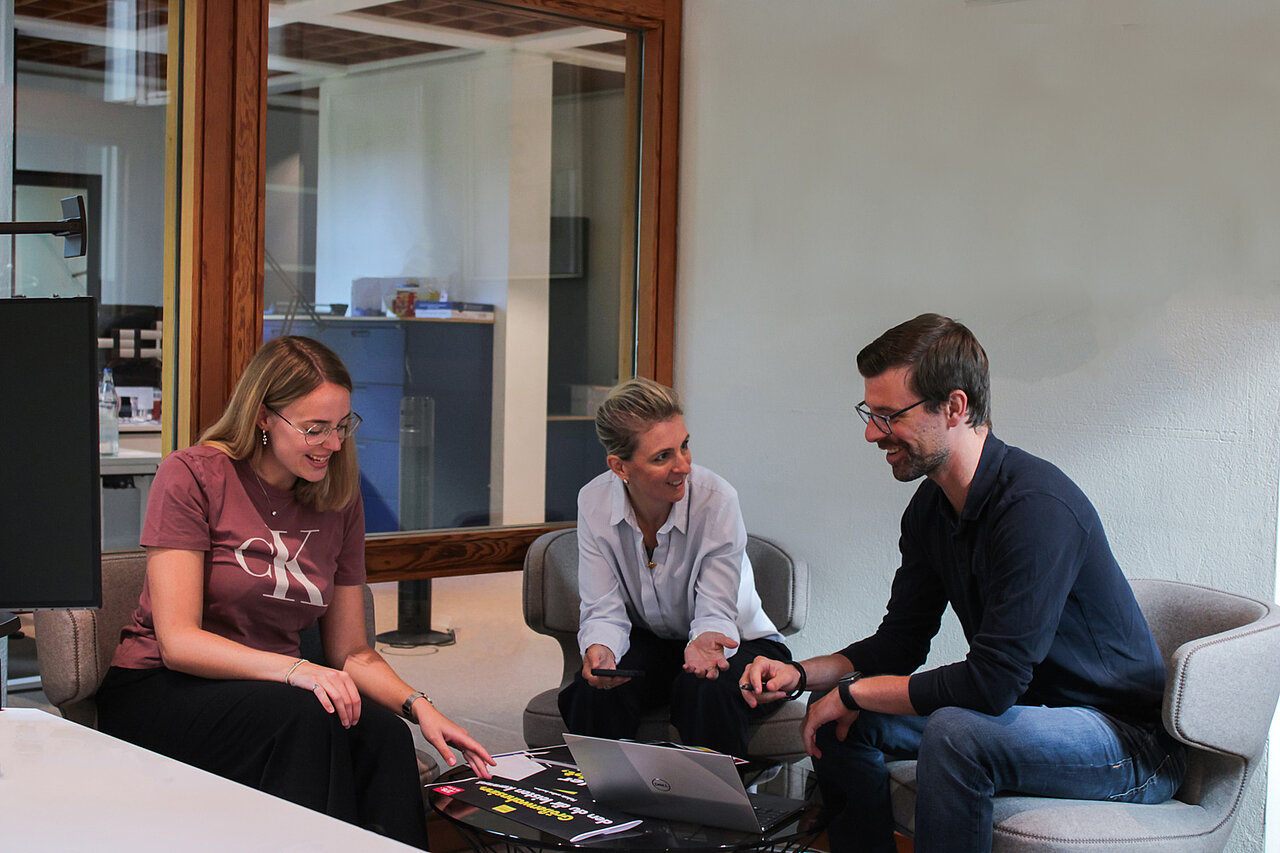 Drei Mitarbeitende von Plakat-verkauft.de sitzen gemeinsam in einem Besprechungsraum, lachen und arbeiten an einem Projekt