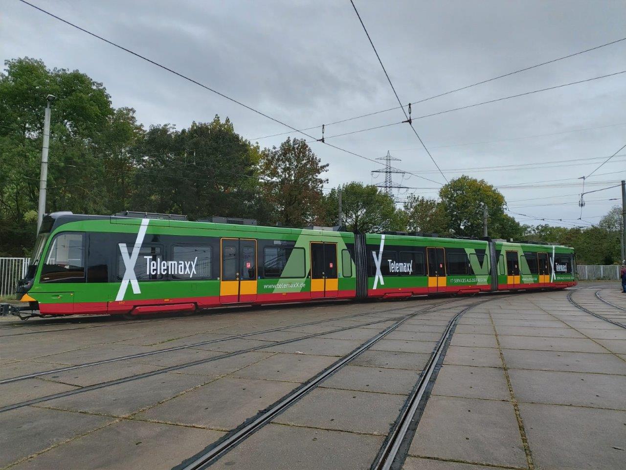 Straßenbahn mit auffälliger TelemaxX-Vollbeklebung in Grün, Rot und Schwarz auf einem Betriebshof, umgeben von Oberleitungen und Bäumen im Hintergrund.